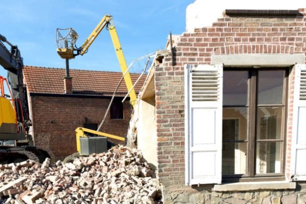 chimney demolition Dismantling a house more than 100 years old. Facade still remaining. Background hydraulic lift and excavator. Front heap of broken bricks laying on ground from demolition